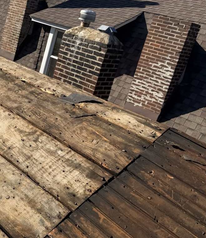 A stone capped brick chimney and a second brick chimney with patches of spalled brick face, standing above a roof stripped back to its plank decking