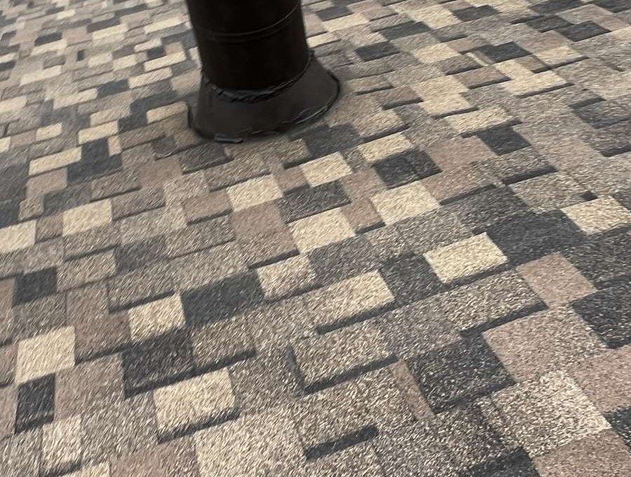 A dark metal flue rising through a shingle roof, with a black flashing collar sealed down onto the shingles around its base